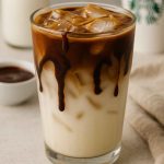 Iced Starbucks-style Coconut Milk Mocha Macchiato with rich espresso drips and coconut milk layers in a clear glass on a neutral countertop.